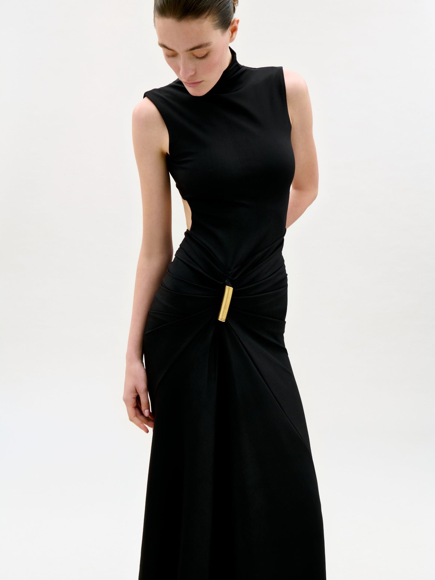 A woman stands against a plain light background, looking down with her hair pulled back, wearing the Bogna Dress—a sleeveless black maxi with a high neckline, gathered waist, gold clasp accent, and asymmetric skirt.