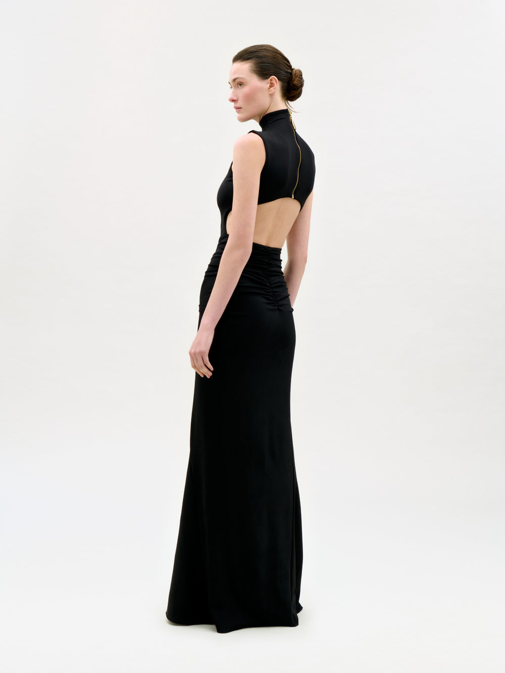 A woman stands in profile wearing the Bogna Dress, a sleek black maxi with an open back and high neckline, crafted from luxurious Italian fabric. Her hair is in a neat bun against a plain light background.