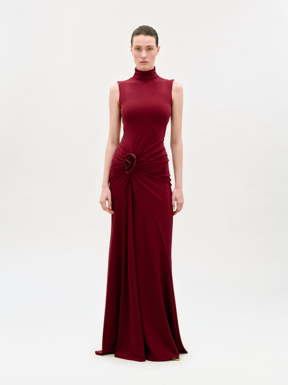 A person faces forward, wearing the Bogna Dress—a sleeveless, burgundy, floor-length dress with a high neckline and gathered waist detail—against a plain, light-colored background.