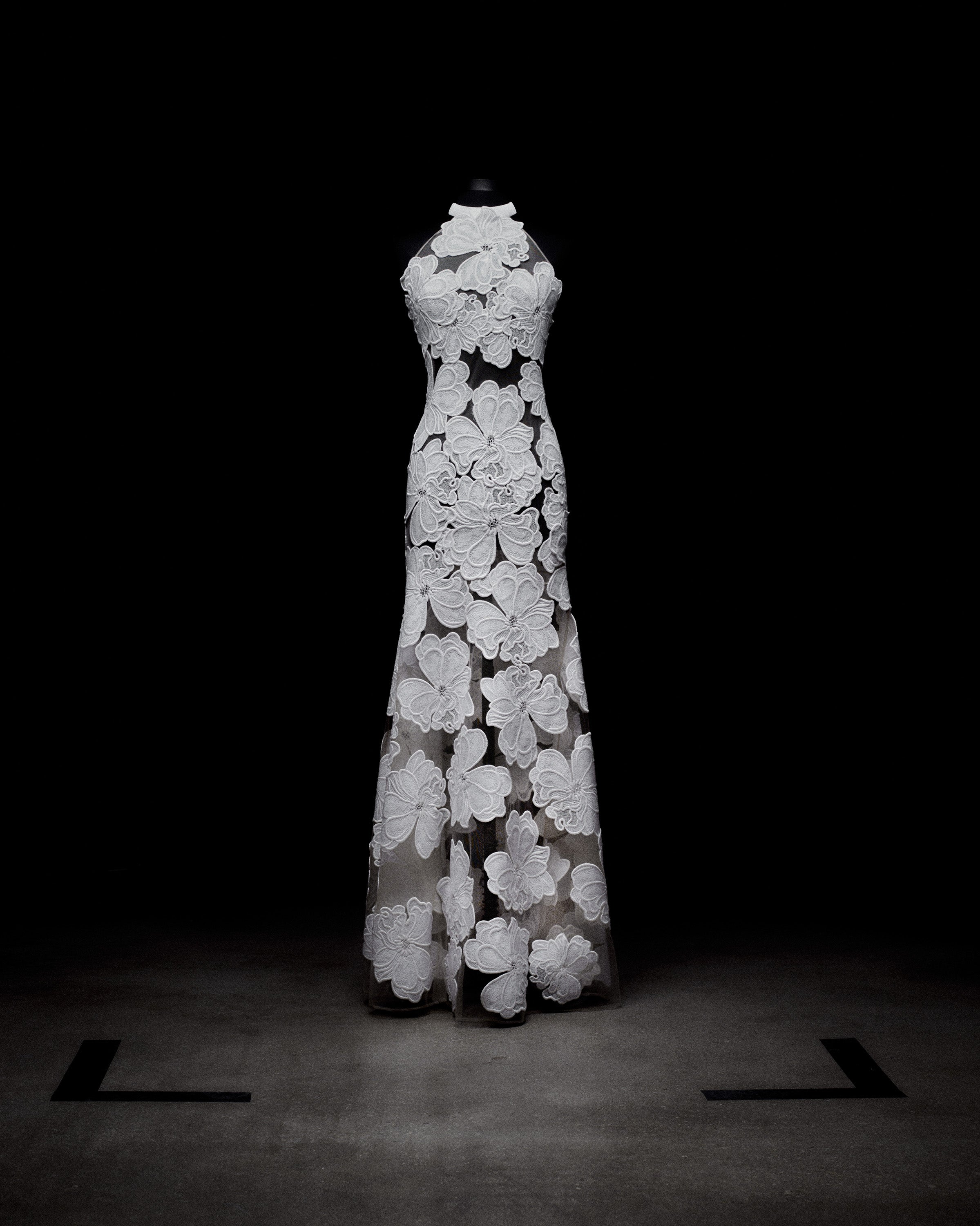 A floor-length dress with a halter neckline, featuring a sheer fabric adorned with large, intricate floral patterns, displayed in a dimly lit space.