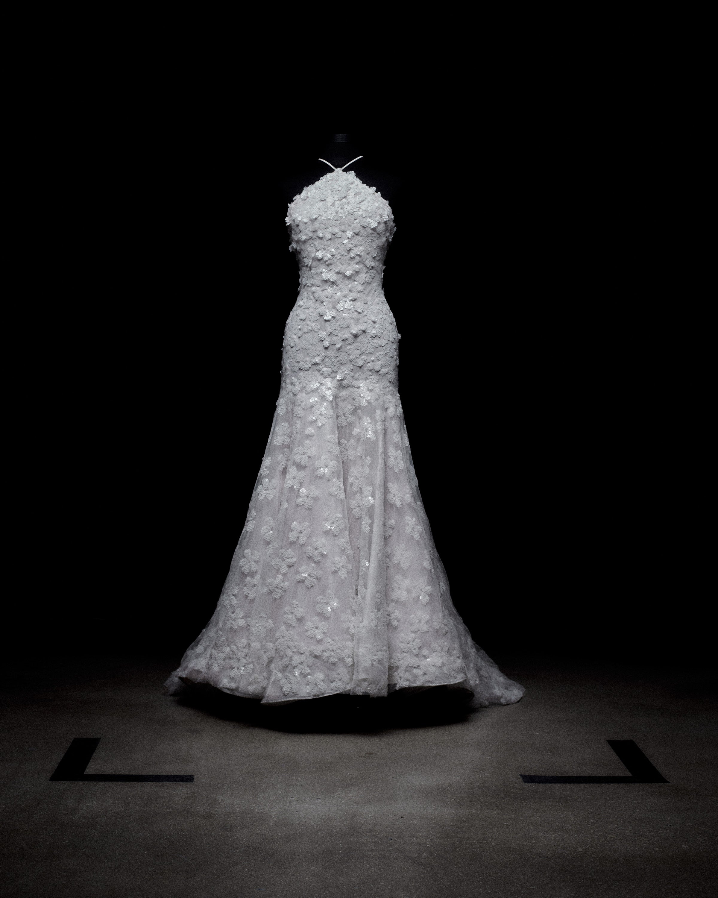 A long, white floral lace gown on a dark mannequin, set against a black background.