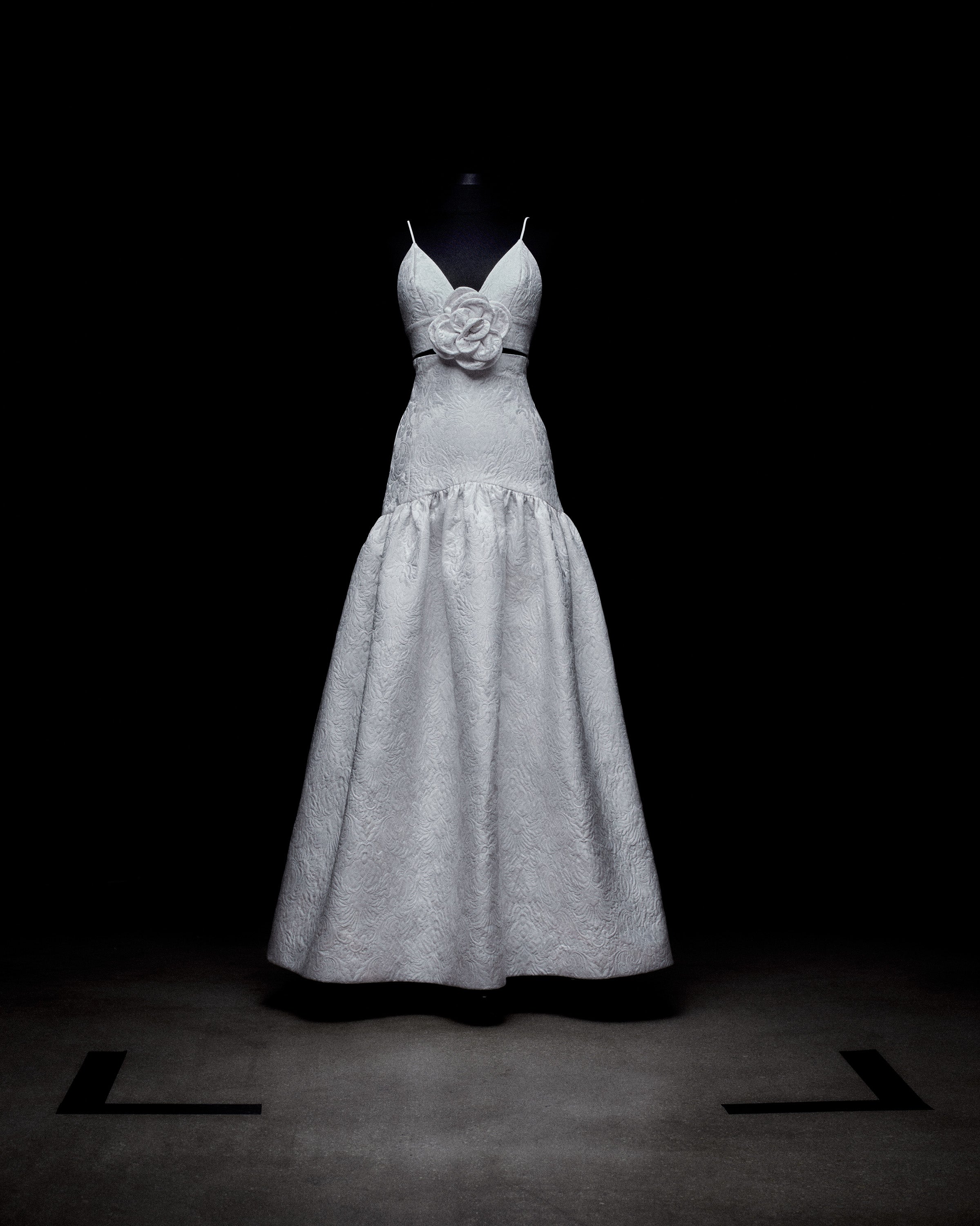 A white sleeveless gown with a fitted bodice, spaghetti straps, a waist flower detail, and a flared skirt on a mannequin against a dark background.