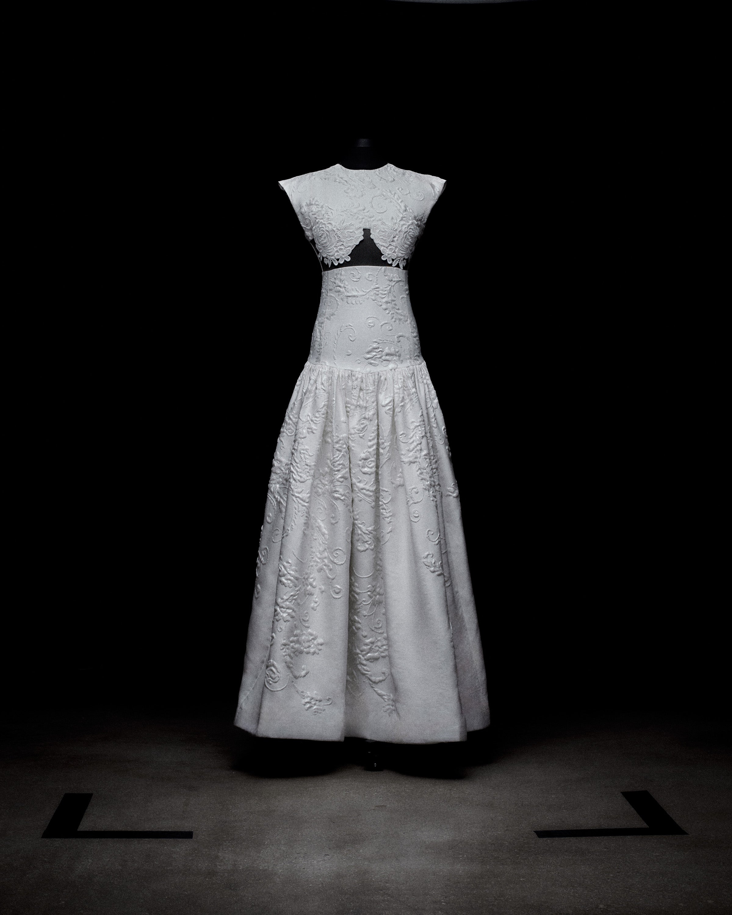 White lace dress on a black mannequin, featuring a sleeveless top with a keyhole cutout and a long skirt decorated with floral patterns, set against a dark background.