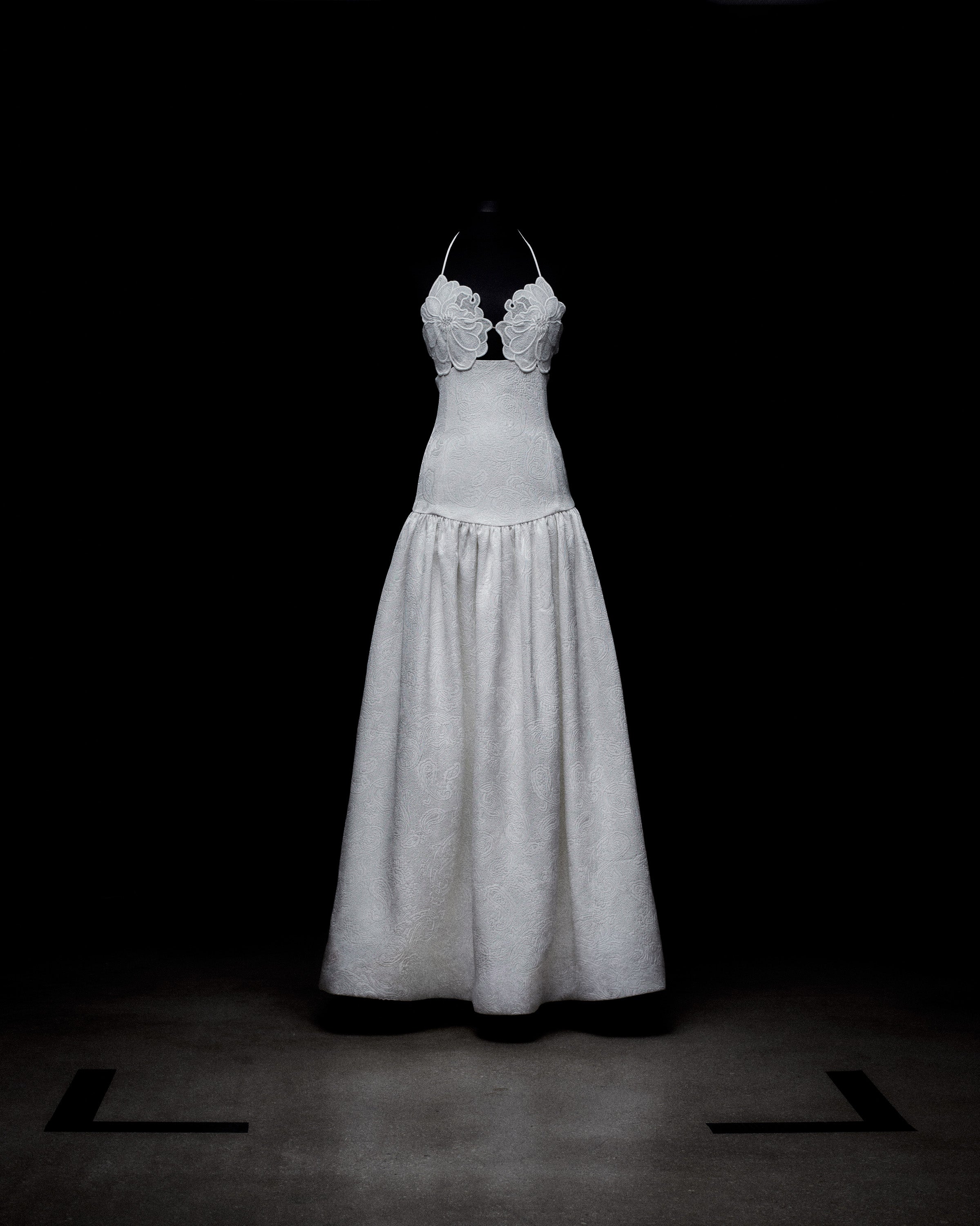 A mannequin displays a white floor-length dress with a fitted bodice and floral detailing on a dark background.