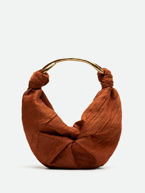 Introducing the Sofia Bag (Small) Bronze, a rust-colored handbag crafted from luxurious Italian fabric with a unique twisted design. It boasts a sleek, gold-toned curved handle that exudes elegance.