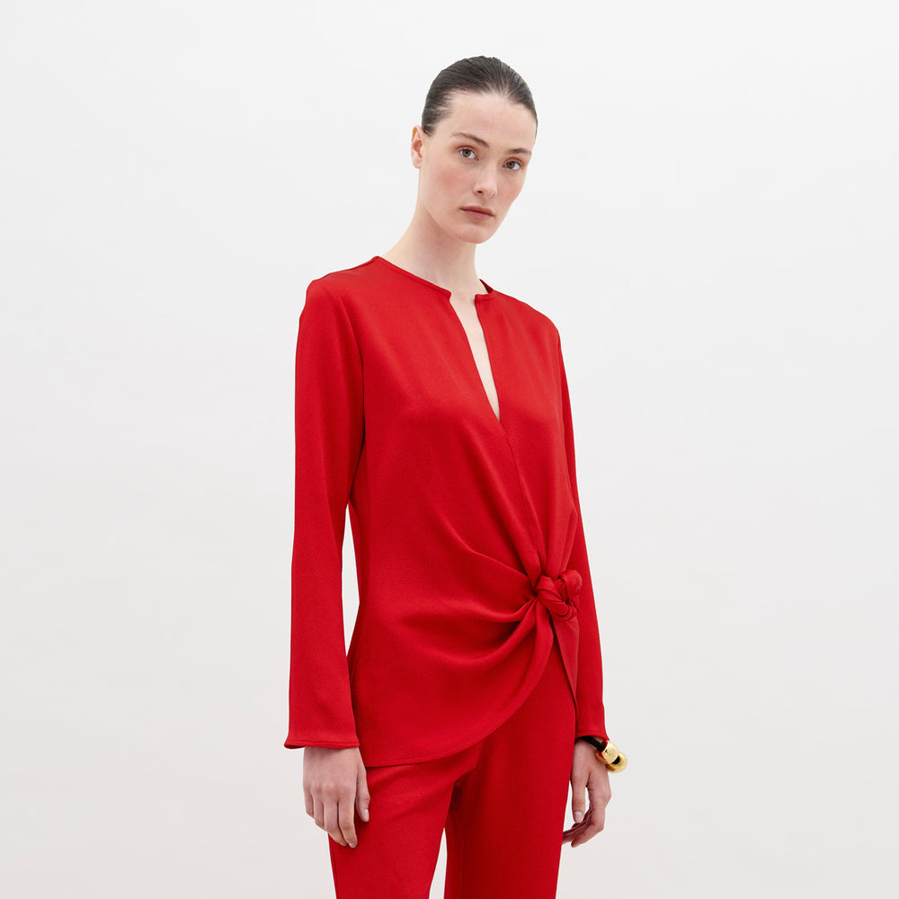 A woman stands in a red outfit with a knot detail on the top. She has her hair pulled back and stares directly into the camera against a plain white background. Available for pre-order now, the Tayra Blouse Rouge will have a ship date of November 15th, 2024.