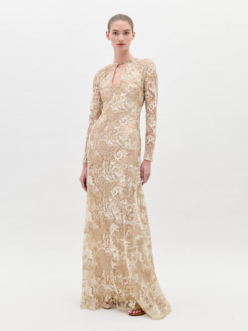 A woman in the Nila Dress Gold, a beige, floor-length, long-sleeved lace gown with a keyhole neckline, stands against a plain background, ready for pre-order ahead of the ship-by date on February 1st, 2025.