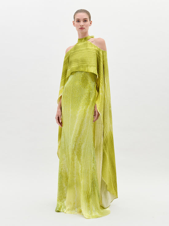 A woman stands wearing the Adelia Dress Green Moire, a long, flowing lime-green gown with off-the-shoulder sleeves and a high neckline. Set against a plain, light-colored background, this stunning dress is available for pre-order now to ensure it reaches you by the ship-by date of February 1st, 2025.