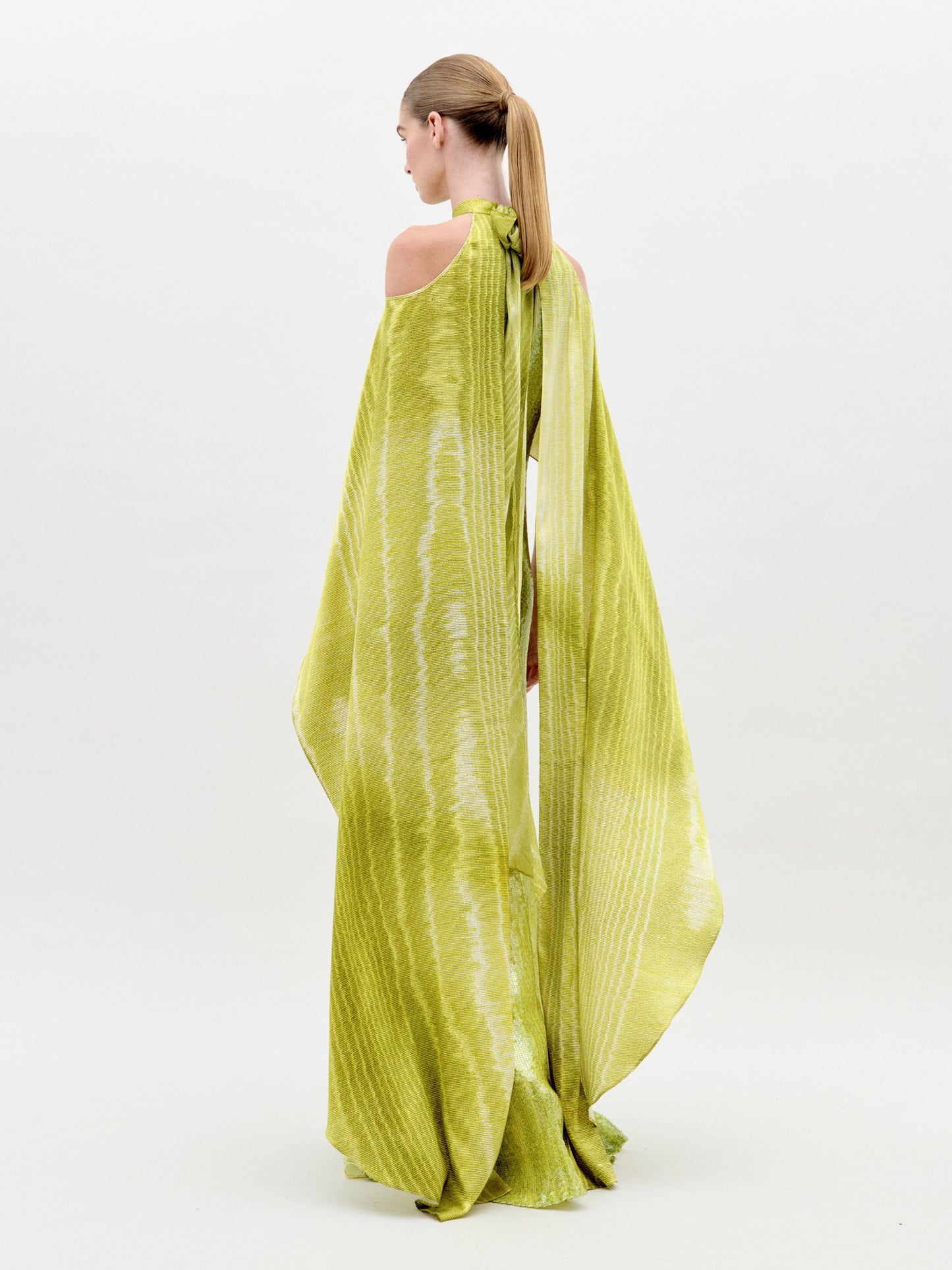 A woman stands wearing the Adelia Dress Green Moire, a long, flowing lime-green gown with off-the-shoulder sleeves and a high neckline. Set against a plain, light-colored background, this stunning dress is available for pre-order now to ensure it reaches you by the ship-by date of February 1st, 2025.