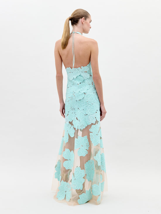 A woman wearing the Mab Dress Turquoise, a light blue floral-patterned halter neck dress with a sheer skirt, stands against a plain background, eagerly awaiting the pre-order ship-by date of February 1st.