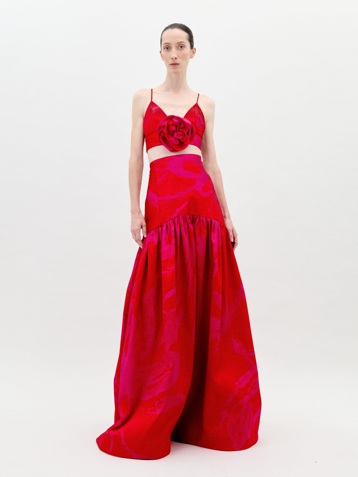 A person is standing wearing the Helen Set Dress Rouge Fuchsia Flower, a red, sleeveless gown featuring a floral detail on the bodice and a long skirt. The background is plain white. This stunning piece will be available for pre-order soon, with an expected ship-by date in early 2025.