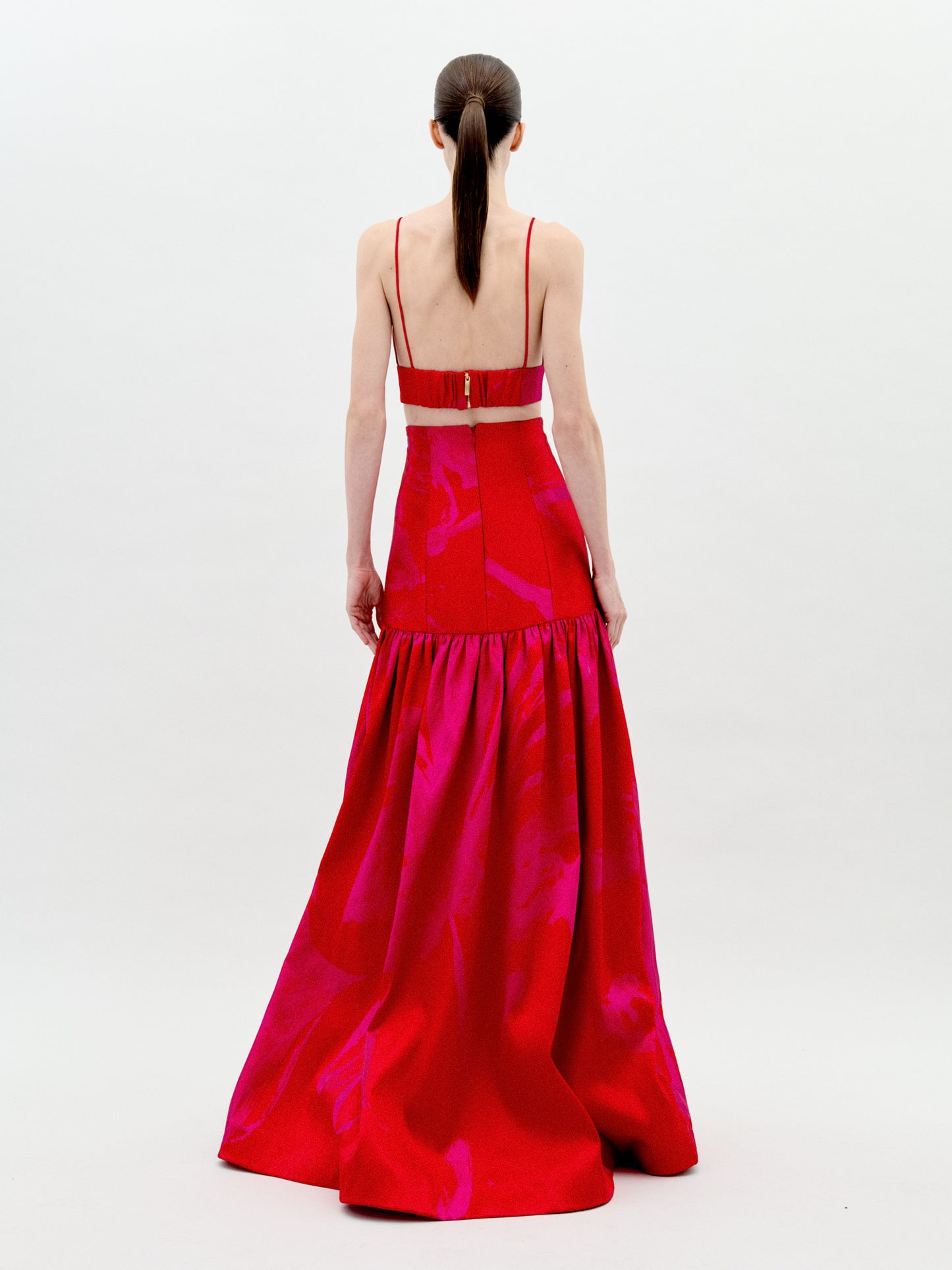 A person is standing wearing the Helen Set Dress Rouge Fuchsia Flower, a red, sleeveless gown featuring a floral detail on the bodice and a long skirt. The background is plain white. This stunning piece will be available for pre-order soon, with an expected ship-by date in early 2025.