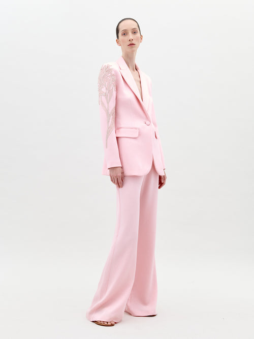 A person stands wearing the Palermo Pant Pink pantsuit, featuring floral detailing on the left shoulder and sleeve, against a plain white background. Available for pre-order now, this elegant outfit will have an exclusive ship-by date of February 1st, 2025.