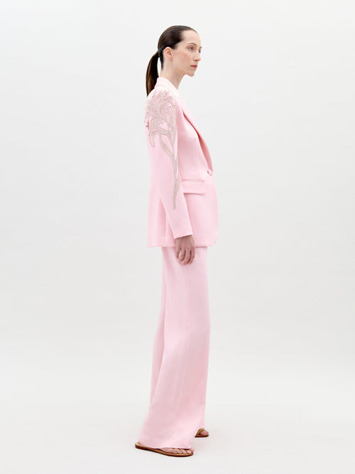 A person stands wearing the Palermo Pant Pink pantsuit, featuring floral detailing on the left shoulder and sleeve, against a plain white background. Available for pre-order now, this elegant outfit will have an exclusive ship-by date of February 1st, 2025.