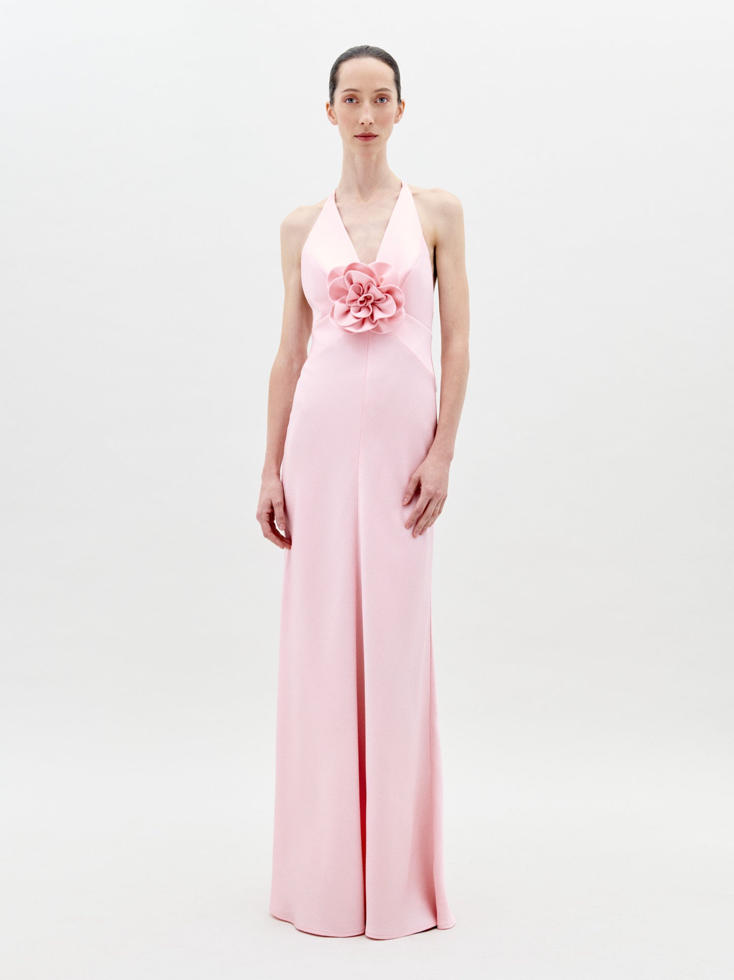 A person stands facing forward wearing the Daniela Dress Pink—a long, pastel pink dress featuring a halter neck and a large rose embellishment at the front. The background is plain white. Available for pre-order now, the dress will ship by February 1st, 2025.