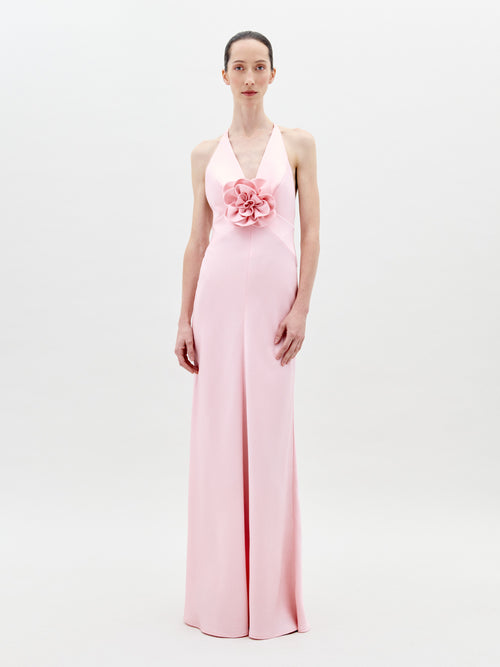 A person stands facing forward wearing the Daniela Dress Pink—a long, pastel pink dress featuring a halter neck and a large rose embellishment at the front. The background is plain white. Available for pre-order now, the dress will ship by February 1st, 2025.