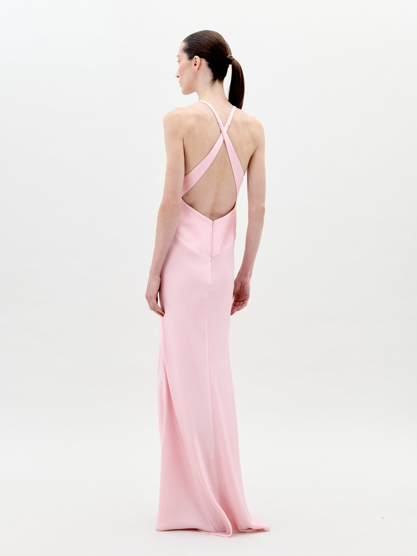 A person stands facing forward wearing the Daniela Dress Pink—a long, pastel pink dress featuring a halter neck and a large rose embellishment at the front. The background is plain white. Available for pre-order now, the dress will ship by February 1st, 2025.