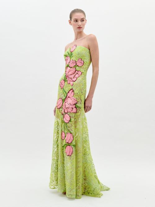 A woman stands wearing the Tulip Dress Rose Tulips, a strapless, green lace dress adorned with large pink floral designs, looking to her left against a plain light background. Pre-order now for exclusive access; shipping by February 1st, 2025.