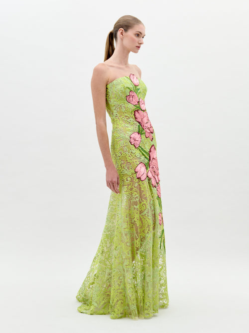 A woman stands wearing the Tulip Dress Rose Tulips, a strapless, green lace dress adorned with large pink floral designs, looking to her left against a plain light background. Pre-order now for exclusive access; shipping by February 1st, 2025.