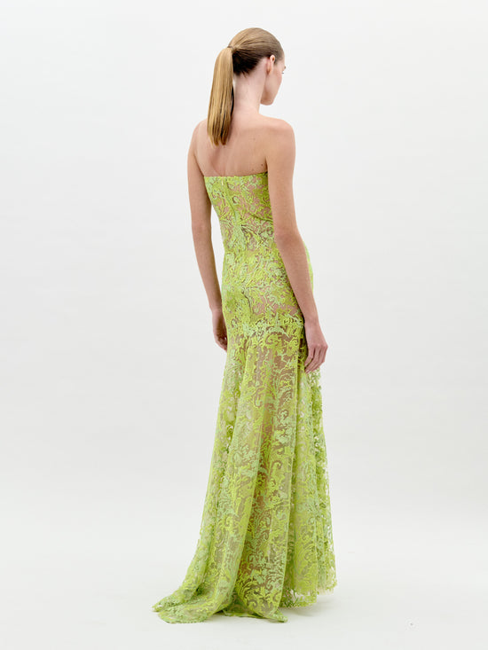 A woman stands wearing the Tulip Dress Rose Tulips, a strapless, green lace dress adorned with large pink floral designs, looking to her left against a plain light background. Pre-order now for exclusive access; shipping by February 1st, 2025.