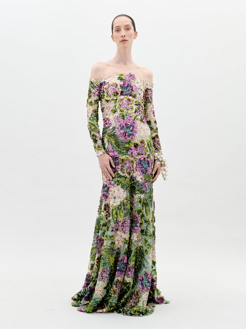 A person stands against a plain background wearing the Rafaella Dress Botanical Sequins, a floor-length, off-the-shoulder floral gown with long sleeves and adorned with a vibrant pattern of flowers and greenery. This elegant piece is available for pre-order now, with a ship-by date set for February 2025.