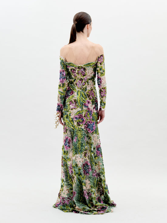 A person stands against a plain background wearing the Rafaella Dress Botanical Sequins, a floor-length, off-the-shoulder floral gown with long sleeves and adorned with a vibrant pattern of flowers and greenery. This elegant piece is available for pre-order now, with a ship-by date set for February 2025.