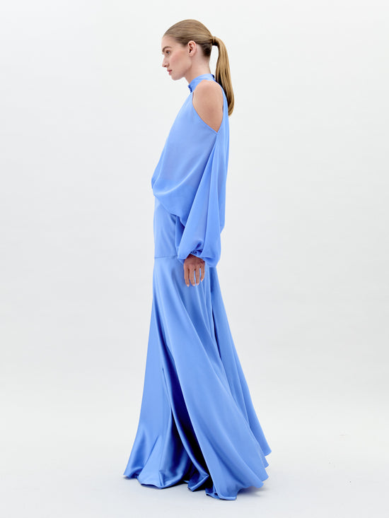 A person in a long, flowing Valeria Dress Sky Blue with shoulder cutouts and a high neckline stands against a plain white background. This elegant attire is available for pre-order now and will ship by February 1st, 2025.