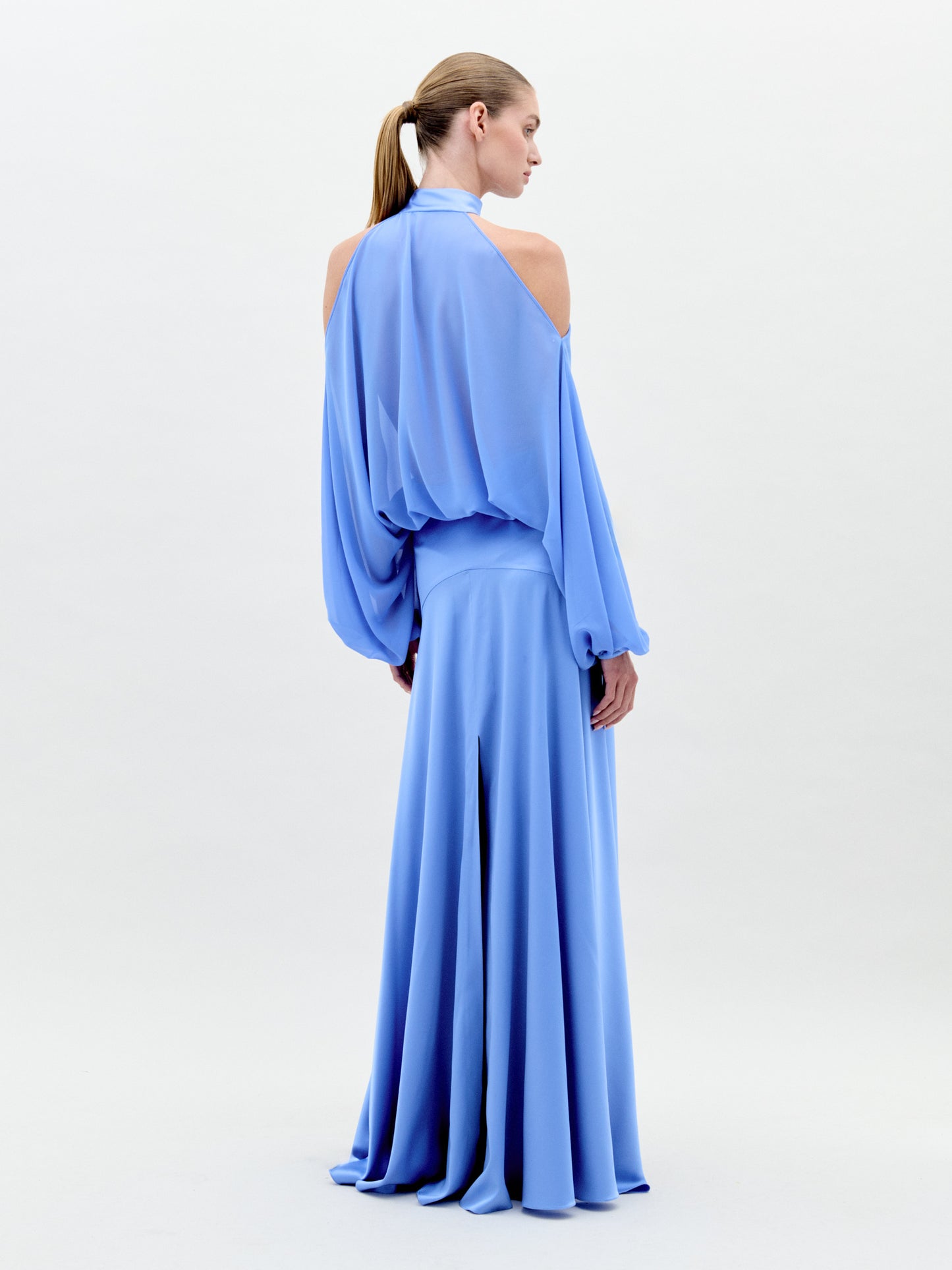 A person in a long, flowing Valeria Dress Sky Blue with shoulder cutouts and a high neckline stands against a plain white background. This elegant attire is available for pre-order now and will ship by February 1st, 2025.