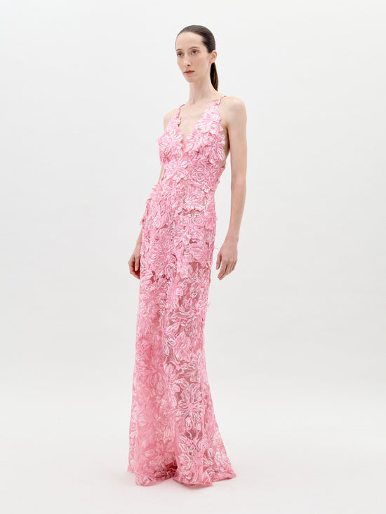 A person stands wearing the Daira Dress Pink, a long dress with thin straps and a beautiful floral pattern. The background is plain and light-colored. This elegant dress is now available for pre-order and will ship by February 1st, 2025.