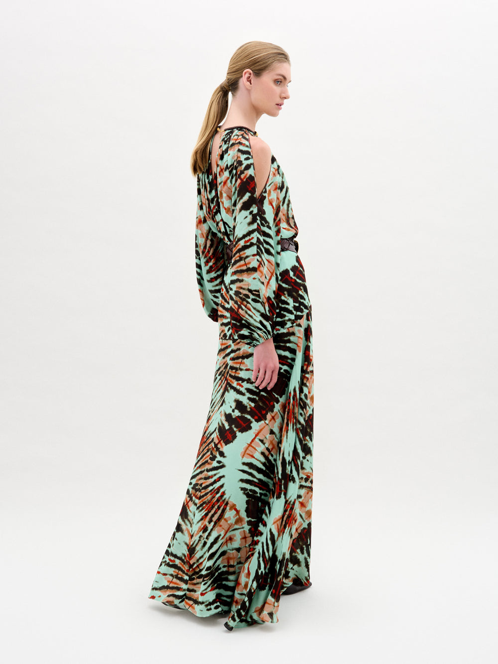 A woman wears the Carla Dress, a long patterned maxi with cut-out shoulders, a gathered neckline, long sleeves, a wide belt, and crafted from luxurious Italian fabric, standing against a plain white background.