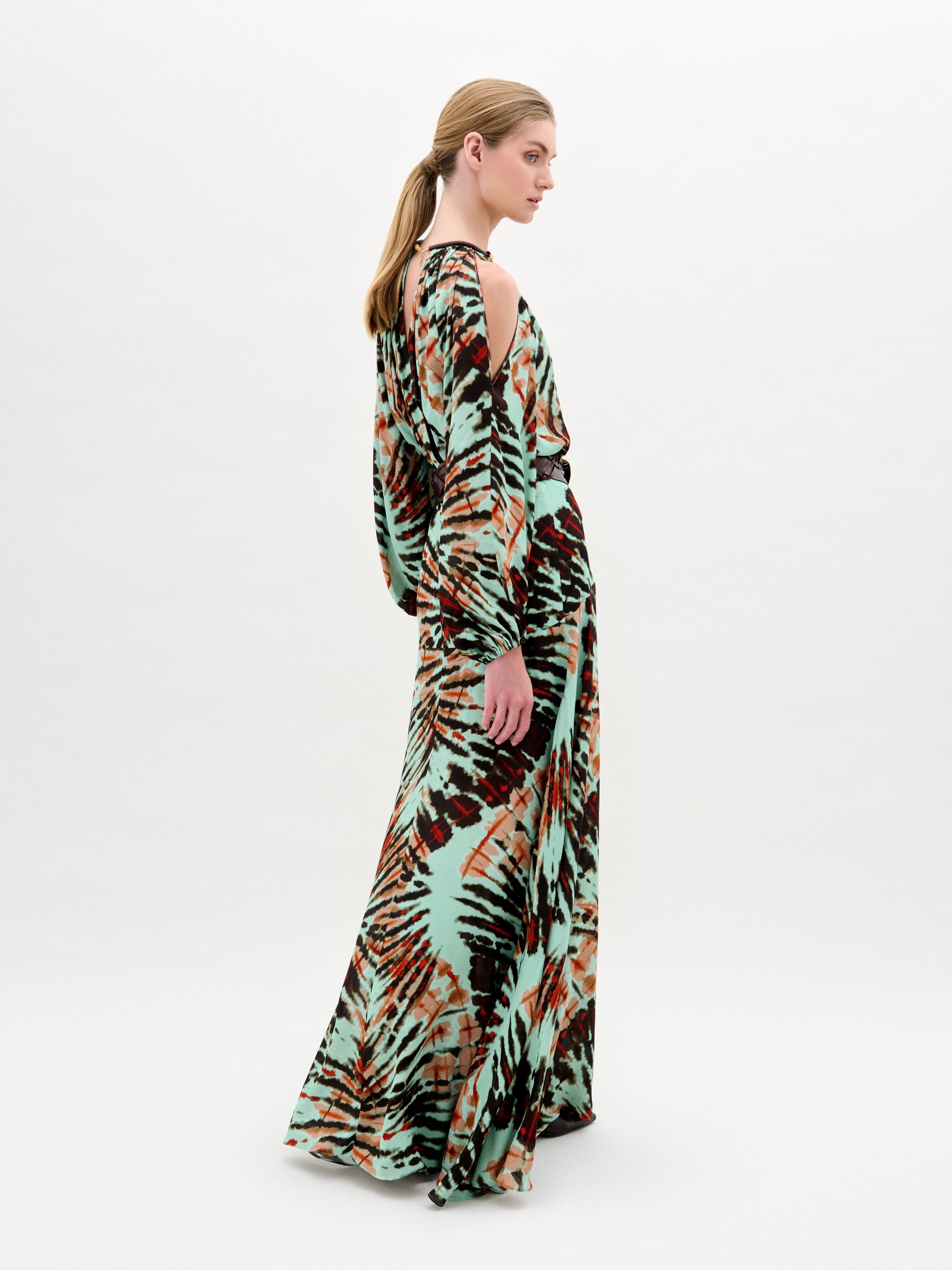 A woman wears the Carla Dress, a long patterned maxi with cut-out shoulders, a gathered neckline, long sleeves, a wide belt, and crafted from luxurious Italian fabric, standing against a plain white background.