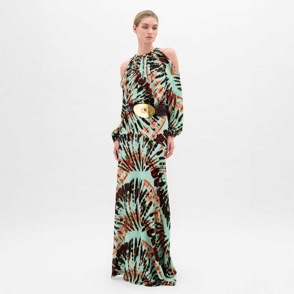 A woman wears the Carla Dress, a long patterned maxi with cut-out shoulders, a gathered neckline, long sleeves, a wide belt, and crafted from luxurious Italian fabric, standing against a plain white background.