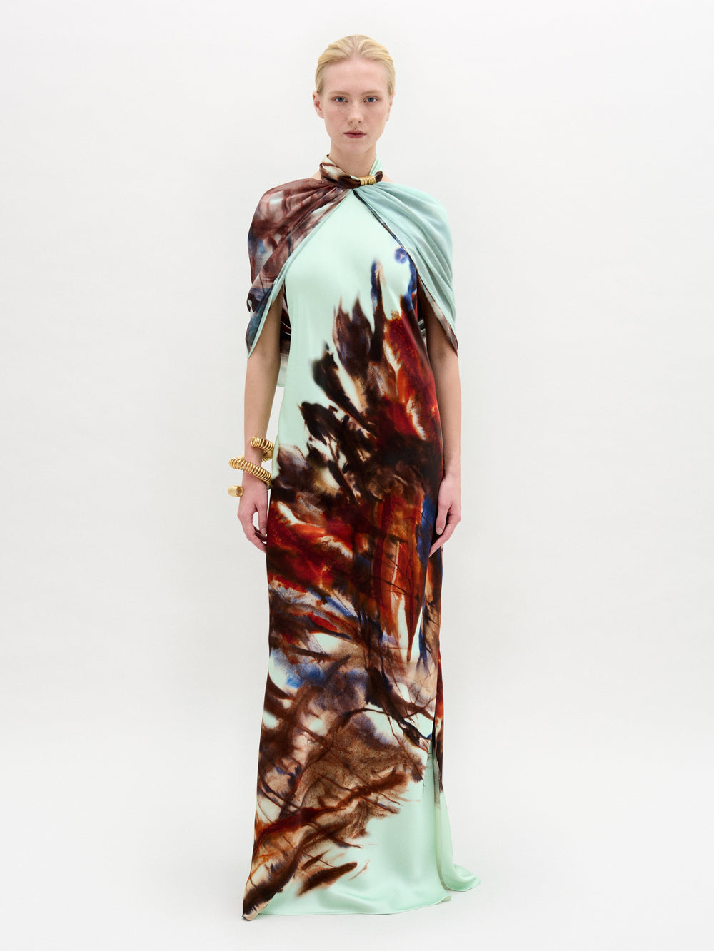 A woman stands against a white background wearing the Carlina Dress, a floor-length light blue maxi made from Italian fabric with a brown and red feather-like pattern and a matching draped shoulder cape.