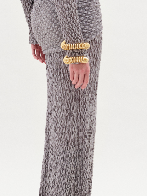 A person faces forward wearing the Ciji Skirt in silver-gray Italian fabric with a textured finish, long sleeves, and a gathered waist, styled with gold bracelets against a white background.