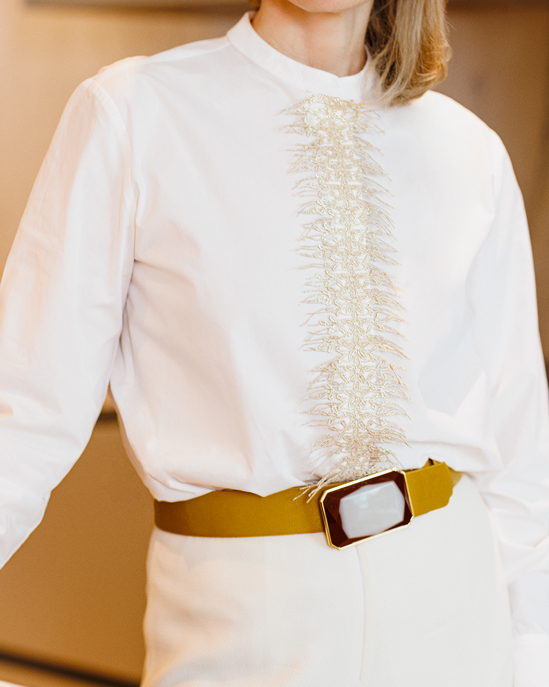 Person wearing a white shirt with intricate, gold embroidery on the front, paired with a mustard-colored belt featuring a large rectangular buckle.