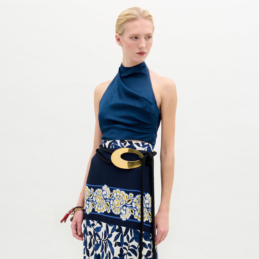 A blonde woman models the Colima Top, a blue high neck crop top, paired with a patterned skirt and large gold belt buckle against a plain white background. This chic outfit is made in Colombia.