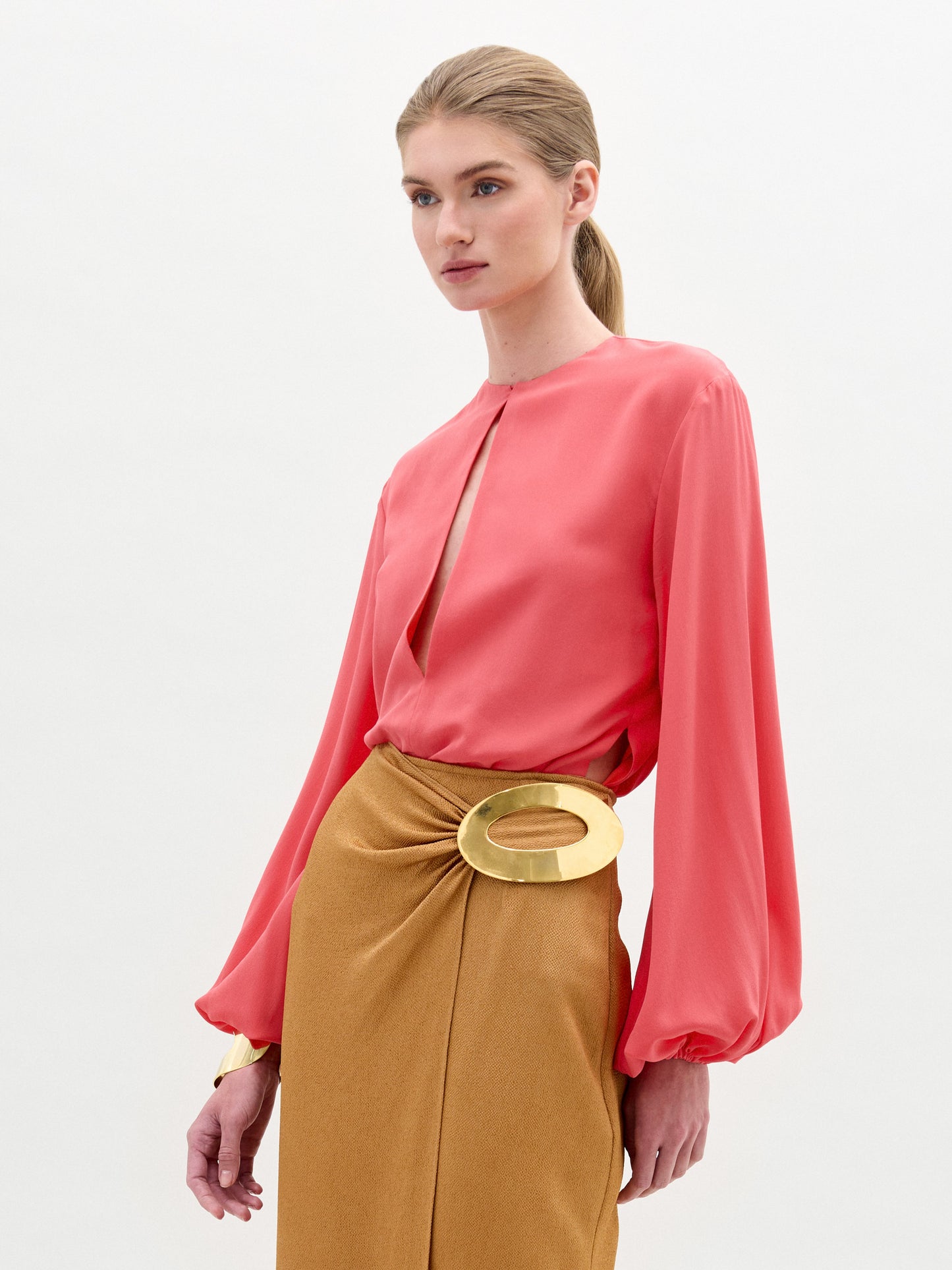 A person wearing the Covilha Blouse, a coral top with a keyhole neckline, and a mustard skirt with a large circular gold buckle, standing against a plain white background.