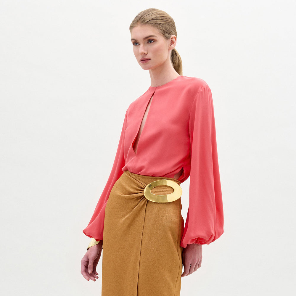 A woman stands against a plain background wearing the Covilha Blouse, a silk coral long-sleeve top, paired with a tan skirt featuring a large gold circular belt detail.