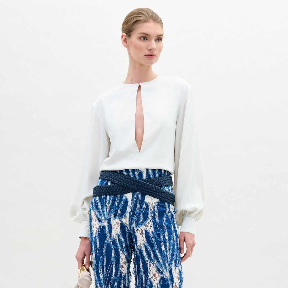A woman poses against a plain background wearing the Covilha Blouse, featuring a keyhole neckline and balloon sleeves, styled with high-waisted blue patterned pants and holding a small beige bag.