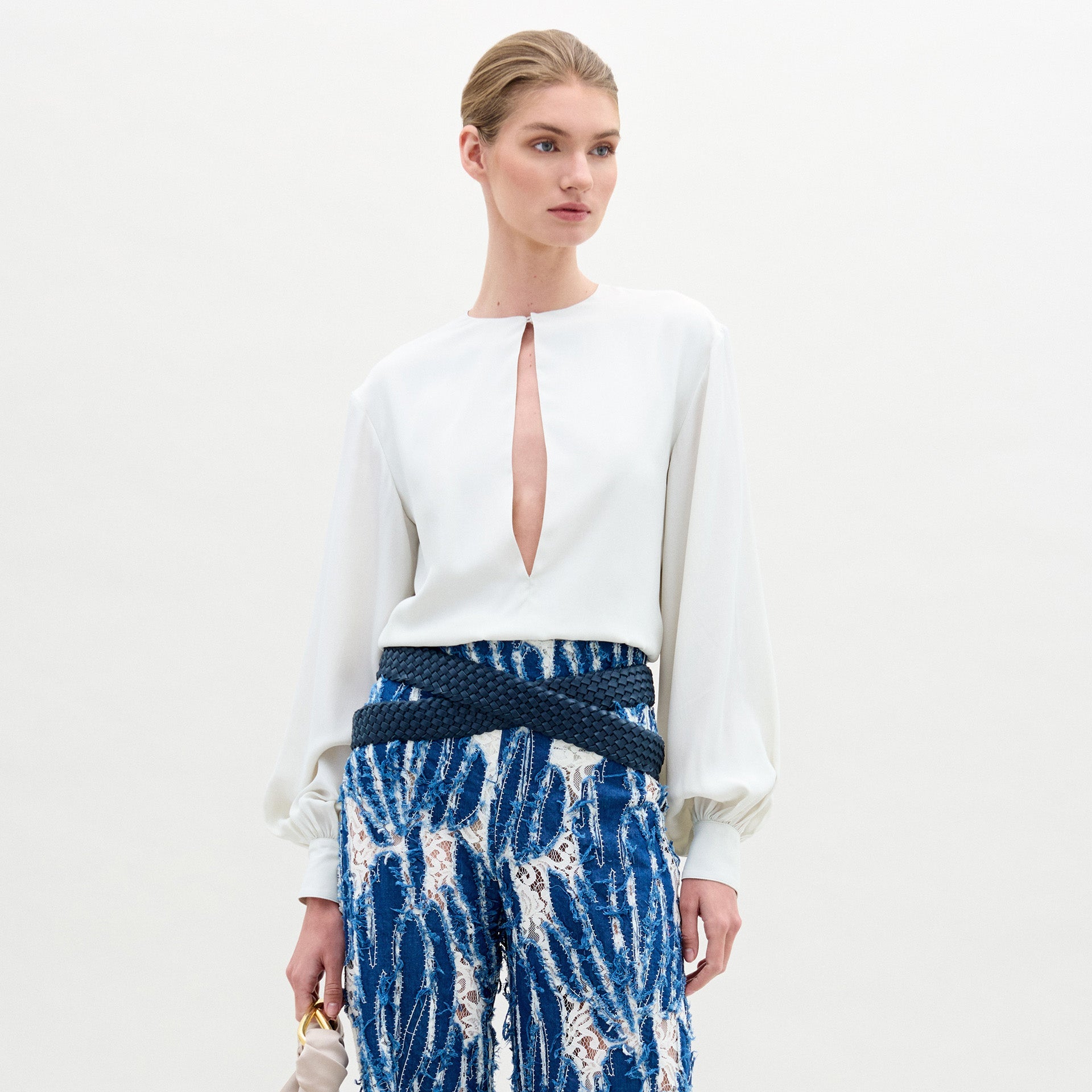 A woman poses against a plain background wearing the Covilha Blouse, featuring a keyhole neckline and balloon sleeves, styled with high-waisted blue patterned pants and holding a small beige bag.