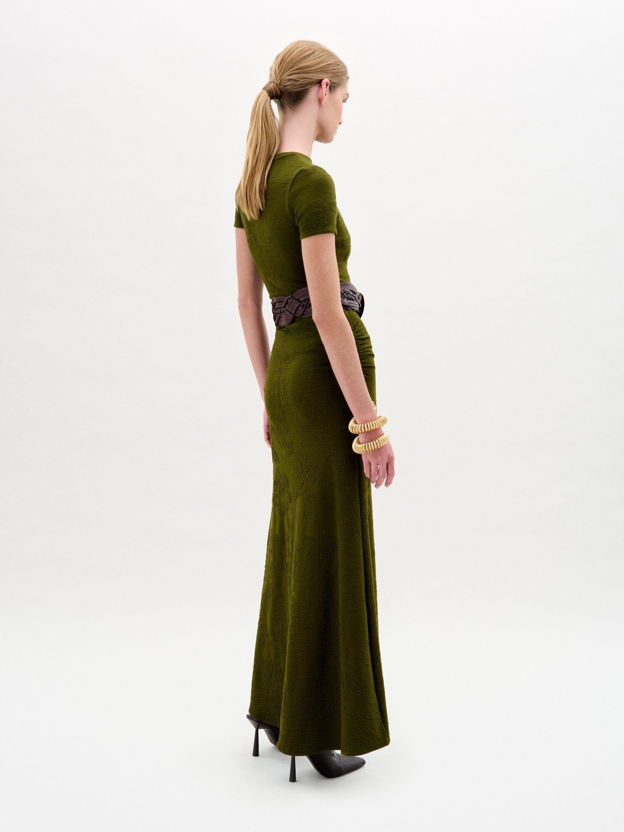 A woman wearing the Cristal Dress, an olive green slim fit maxi dress paired with a wide black belt with a gold buckle, black boots, and gold bracelets stands against a plain white background.