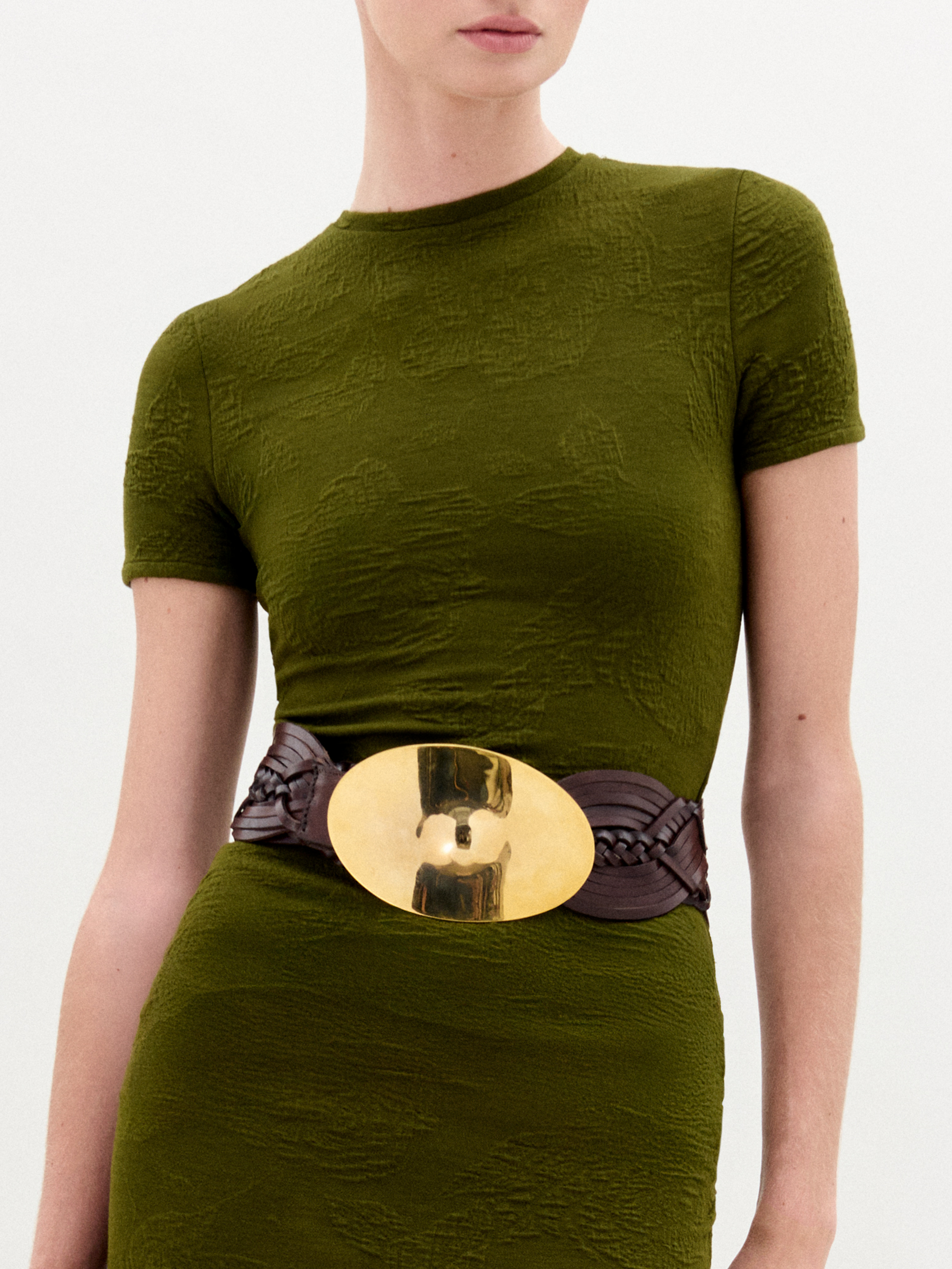 A woman wearing the Cristal Dress, an olive green slim fit maxi dress paired with a wide black belt with a gold buckle, black boots, and gold bracelets stands against a plain white background.