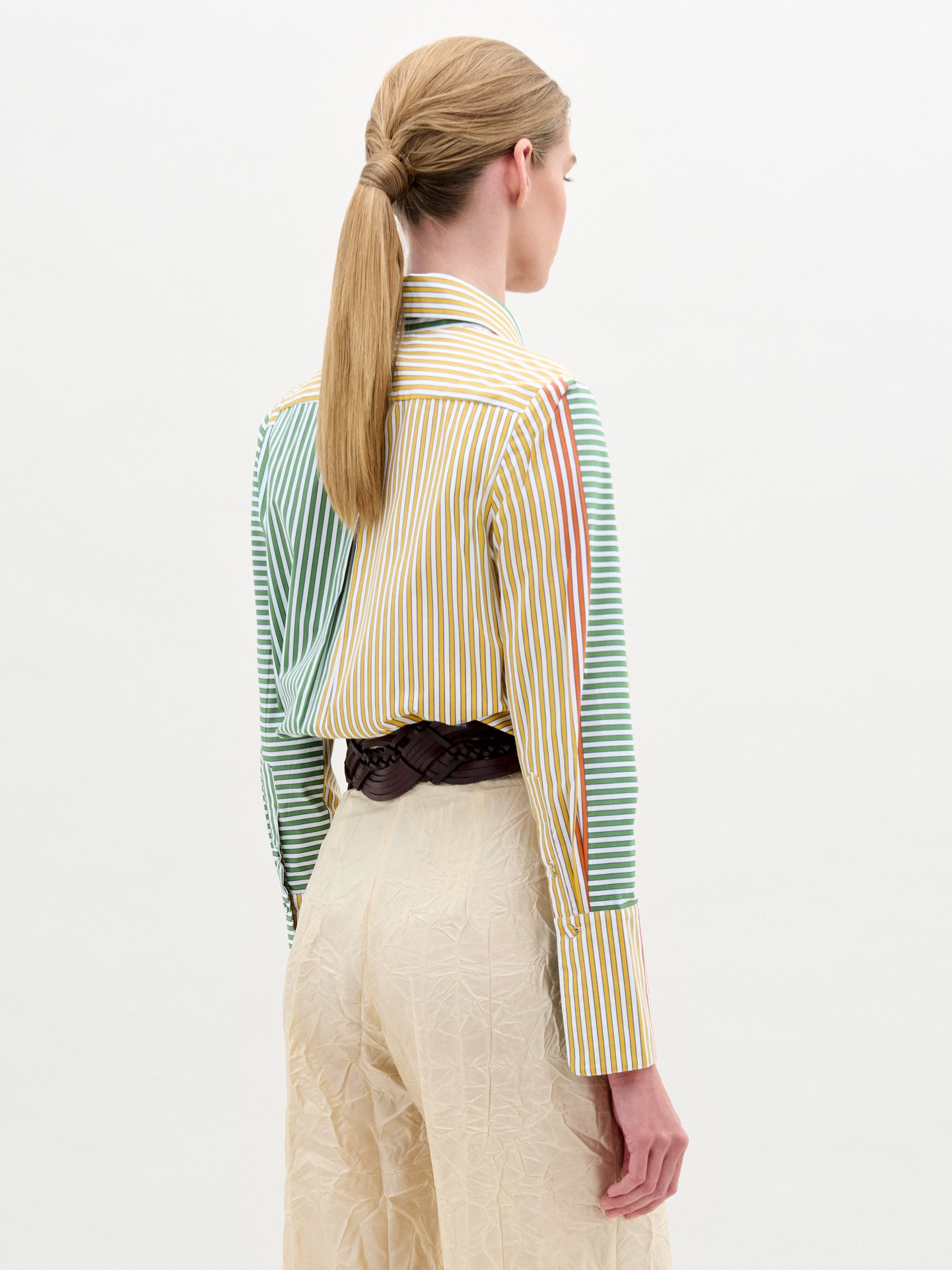 A woman stands against a plain background, wearing the Cubillos Shirt White, a 100% cotton multicolored striped shirt, paired with textured beige pants and a wide belt featuring a large round gold buckle.