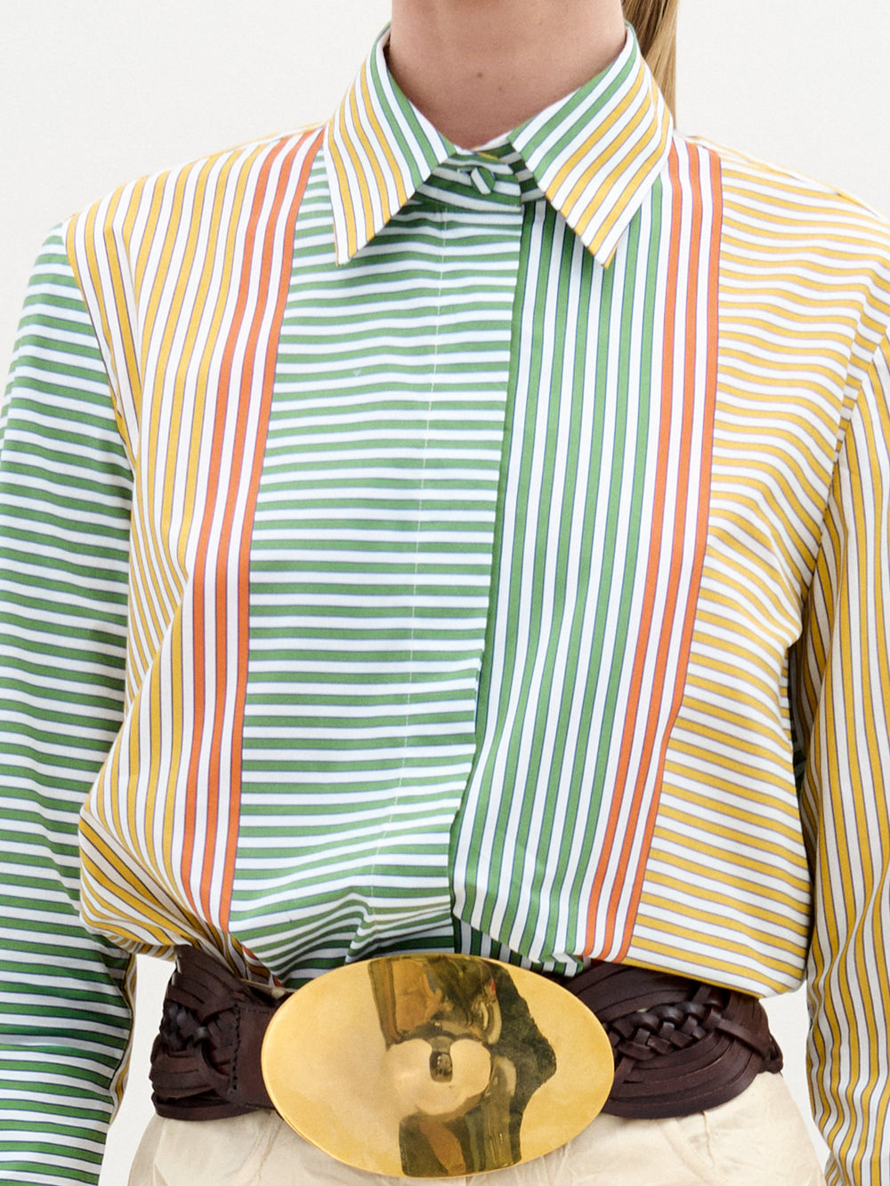 A woman stands against a plain background wearing the Cubillos Shirt, a multicolored 100% cotton striped top, paired with beige textured pants and a large gold belt buckle.