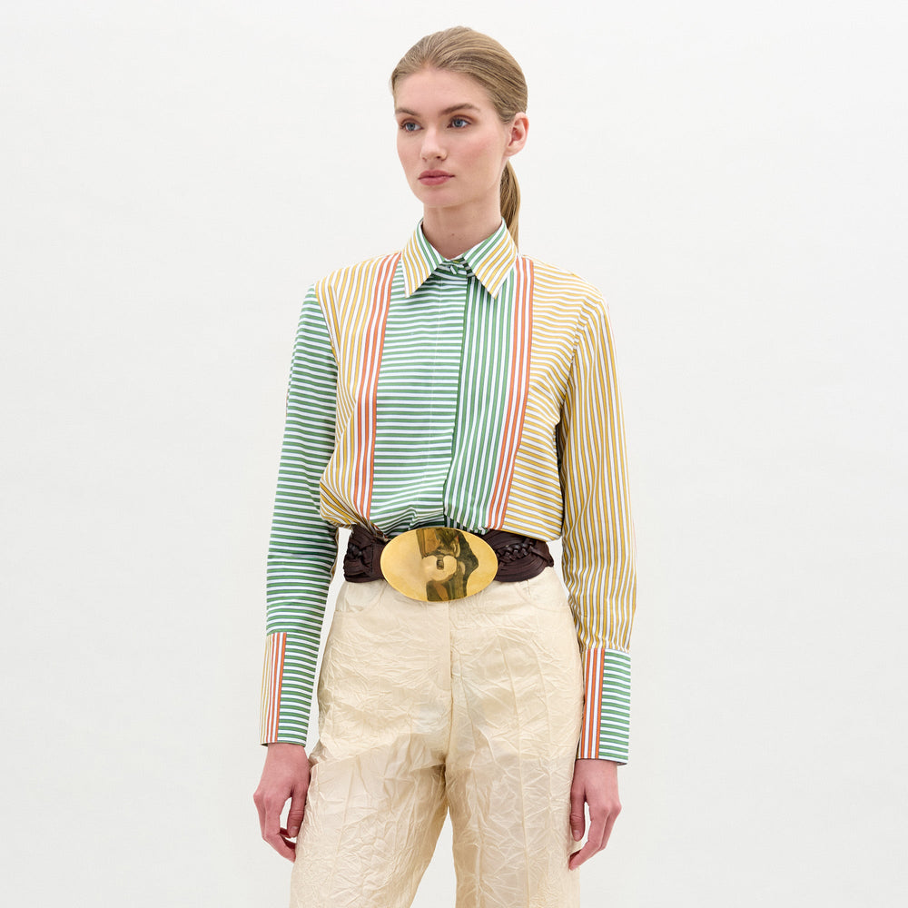 A woman stands against a plain background, wearing the Cubillos Shirt White made from 100% cotton Italian fabric, paired with beige textured pants and a large gold belt buckle.