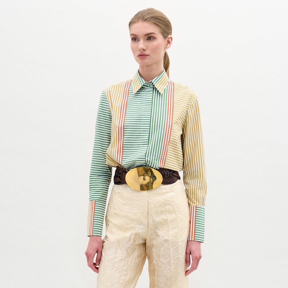 A woman stands against a plain background, wearing the Cubillos Shirt White, a 100% cotton multicolored striped shirt, paired with textured beige pants and a wide belt featuring a large round gold buckle.