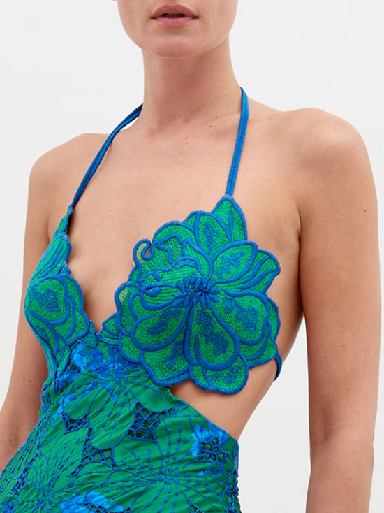 A person stands wearing a vibrant green and blue floral-patterned dress with an open back and a halter neck, posing against a plain white background. The Cara Dress Green Navy Embroidery is available for pre-order now, with a ship by date of November 15th, 2024.