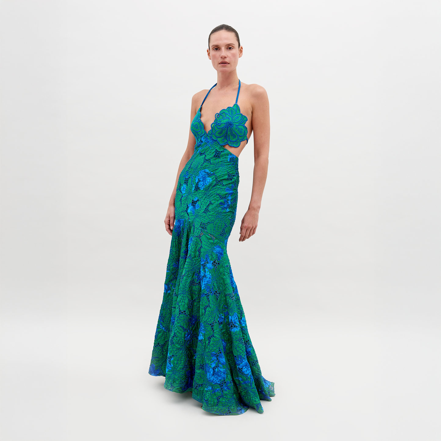 A person stands wearing a vibrant green and blue floral-patterned dress with an open back and a halter neck, posing against a plain white background. The Cara Dress Green Navy Embroidery is available for pre-order now, with a ship by date of November 15th, 2024.