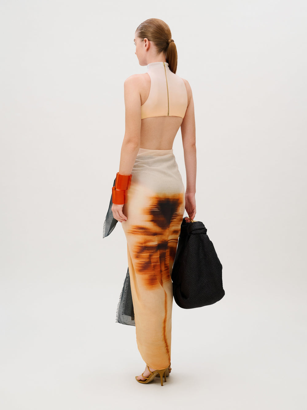 A person seen from behind wears the Caribbean Pareo, a high-neck crop top and a long, sheer skirt with a sunburst-like print in Italian fabric, while carrying a black tote bag.