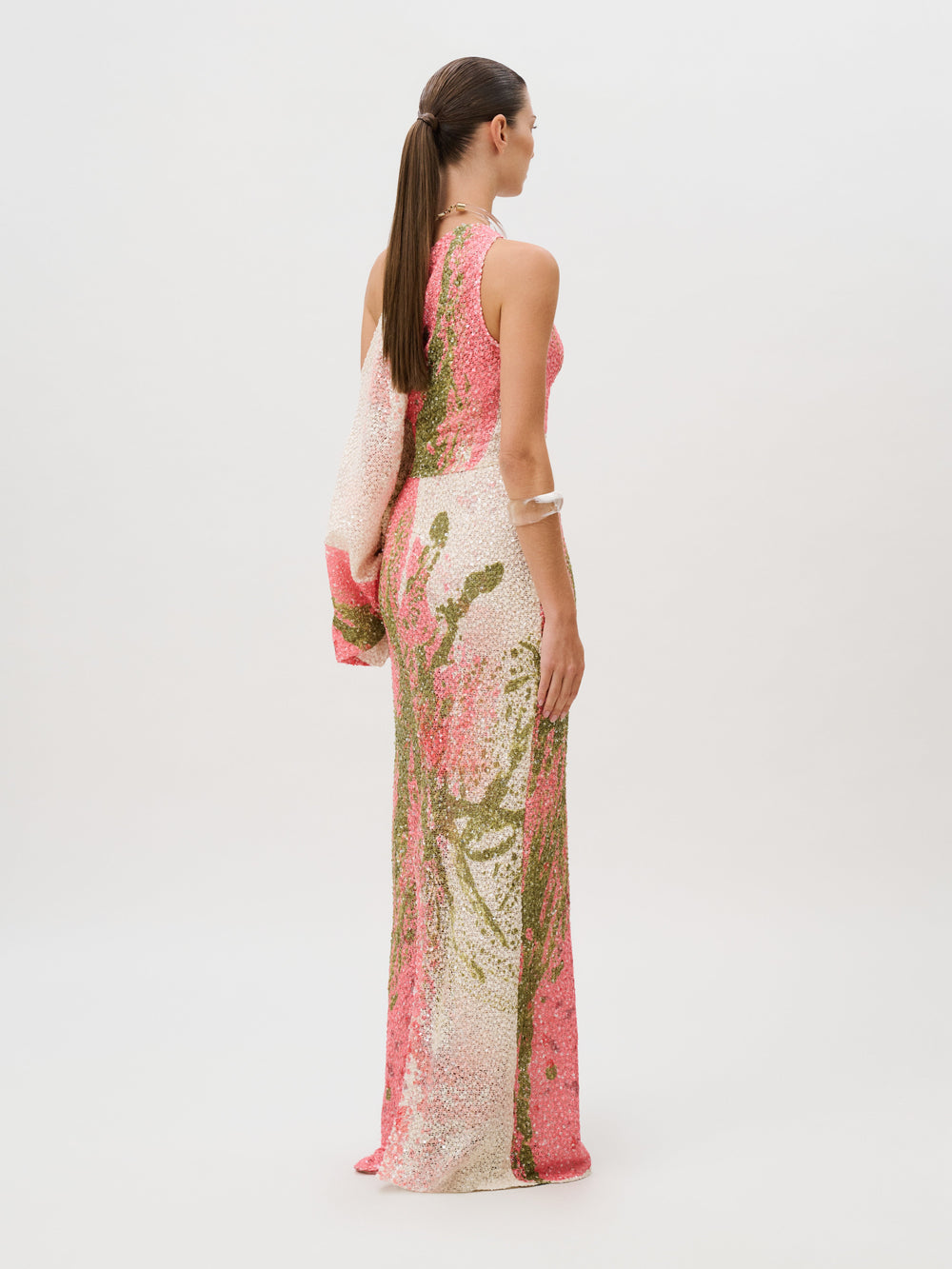 The Carmel Dress is a sleeveless, floor-length sequin gown featuring a pink and green floral pattern crafted from Italian fabric with a single draped sleeve.